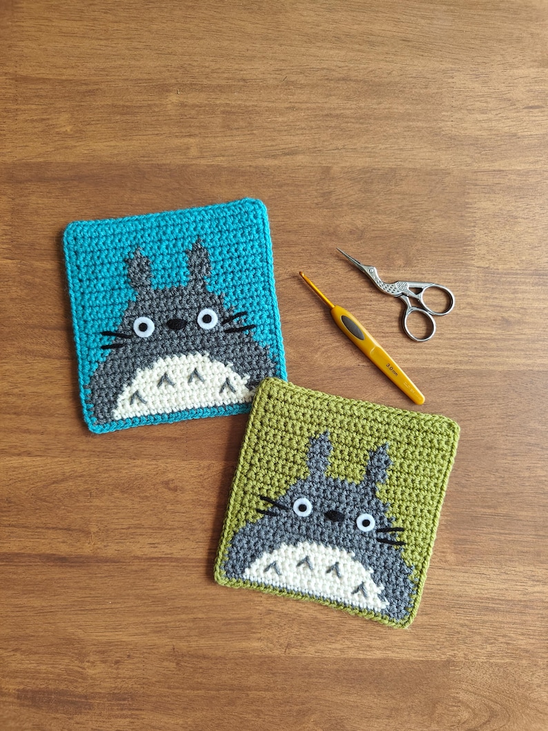 May include: Two crocheted coasters featuring a cartoon character design. One coaster is teal, the other is olive green. Both have a gray character with white accents and black whiskers. A yellow crochet hook and small scissors are also on the wooden surface.