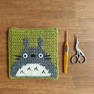 May include: A handmade crochet square featuring a Totoro design in gray, white, and green. A yellow crochet hook and small silver scissors are next to the square on a wooden surface. The square is approximately 4 inches by 4 inches.