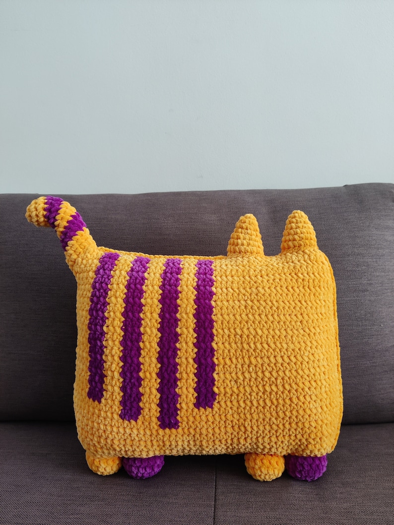 May include: A handmade, cat-shaped cushion in yellow with purple stripes, tail, and feet. The ears are yellow. The cushion is made from a soft, textured material. The design is intended for decorative purposes, adding a playful touch to any room.