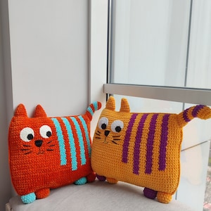 May include: Two handmade crochet cat cushions. One is orange with blue stripes, the other is yellow with purple stripes. Both have large eyes, small ears, and tails. They are on a grey surface near a window.