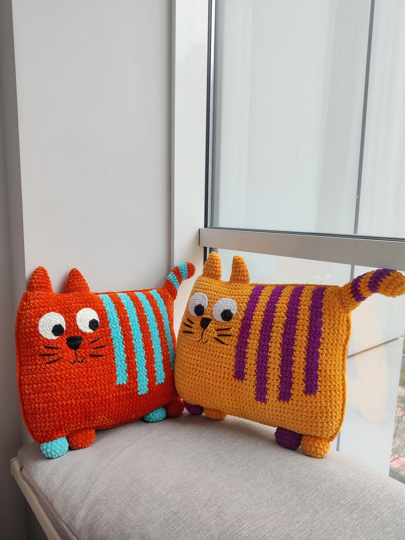May include: Two handmade crochet cat cushions. One is orange with blue stripes, the other is yellow with purple stripes. Both have large eyes, small ears, and tails. They are on a grey surface near a window.