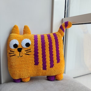 May include: A handmade, plush cat-shaped cushion. The cushion is primarily orange with vertical purple stripes. It has large white eyes, a black nose and whiskers, and small purple feet. The cat has a curved tail and small ears.