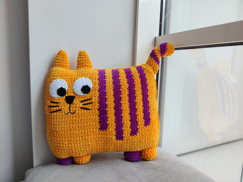 May include: A handmade, plush cat-shaped cushion. The cushion is primarily orange with vertical purple stripes. It has large white eyes, a black nose and whiskers, and small purple feet. The cat has a curved tail and small ears.