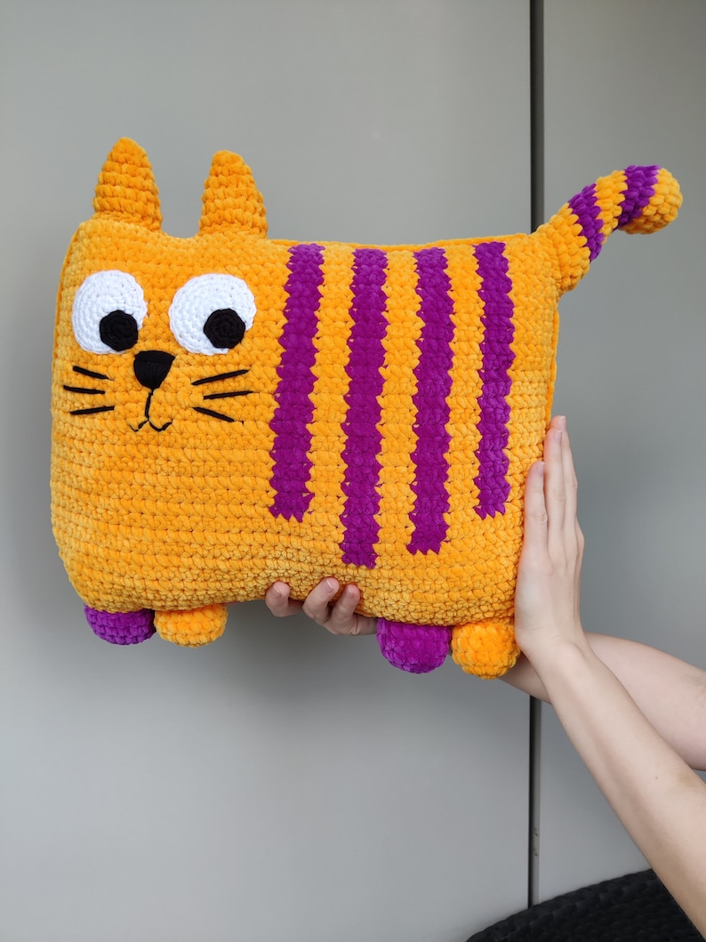 May include: A vibrant, orange cat-shaped cushion with purple stripes. The cushion features large white eyes with black pupils, a black nose, and whiskers. The cat has small ears and a tail with purple stripes. The cushion is being held by two hands.