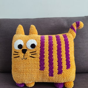 May include: A handmade, plush cat cushion in a rectangular shape. The cushion is primarily orange with vertical purple stripes. It has white eyes, black whiskers, and small purple feet. The cat has a tail that is striped in purple and orange.