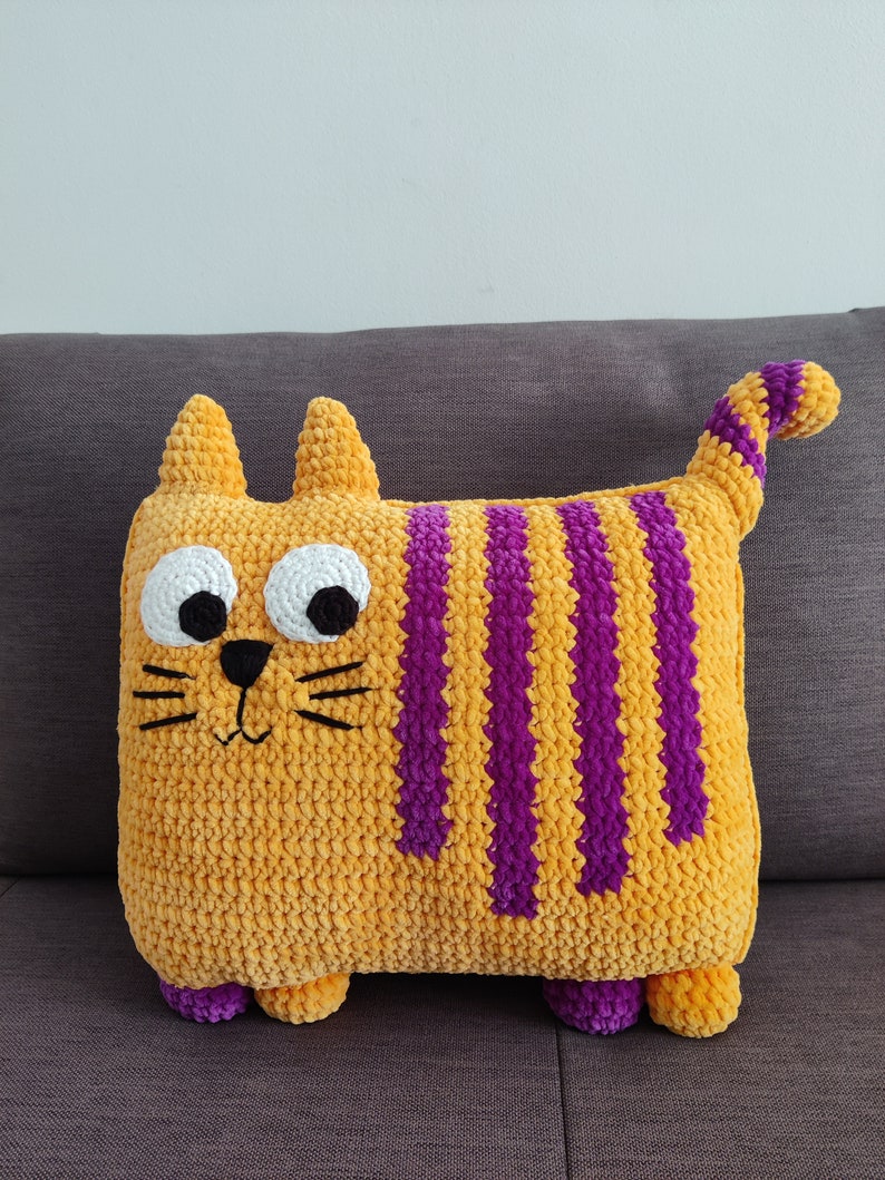 May include: A handmade, plush cat cushion in a rectangular shape. The cushion is primarily orange with vertical purple stripes. It has white eyes, black whiskers, and small purple feet. The cat has a tail that is striped in purple and orange.