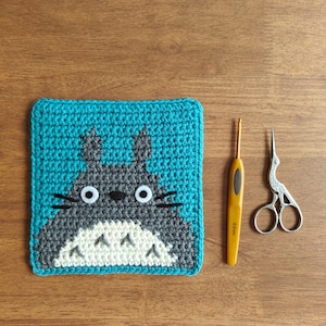 May include: A handmade crochet coaster featuring a cartoon character with a gray body, white belly, and black whiskers, set against a teal background. A yellow crochet hook and small silver scissors are next to the coaster on a wooden surface.