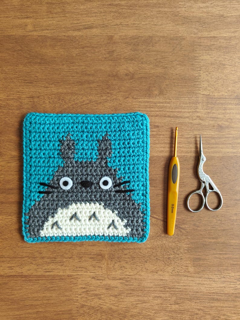 May include: A handmade crochet coaster featuring a cartoon character with a gray body, white belly, and black whiskers, set against a teal background. A yellow crochet hook and small silver scissors are next to the coaster on a wooden surface.