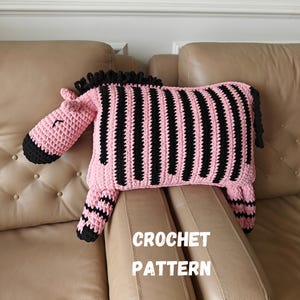 Peut inclure: Un coussin au crochet fait main en forme de zèbre avec un corps rayé rose et noir, une crinière noire et une queue noire. Le coussin présente une tête rose avec des accents noirs et le texte CROCHET PATTERN imprimé en blanc sur la partie inférieure.