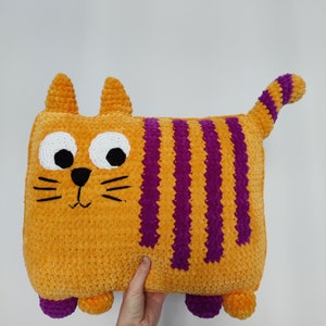 May include: A handmade, orange cat-shaped cushion with purple stripes, white eyes, and a black nose and whiskers. The plush cushion has small orange ears, purple feet, and a striped tail. The cushion is held against a white background.