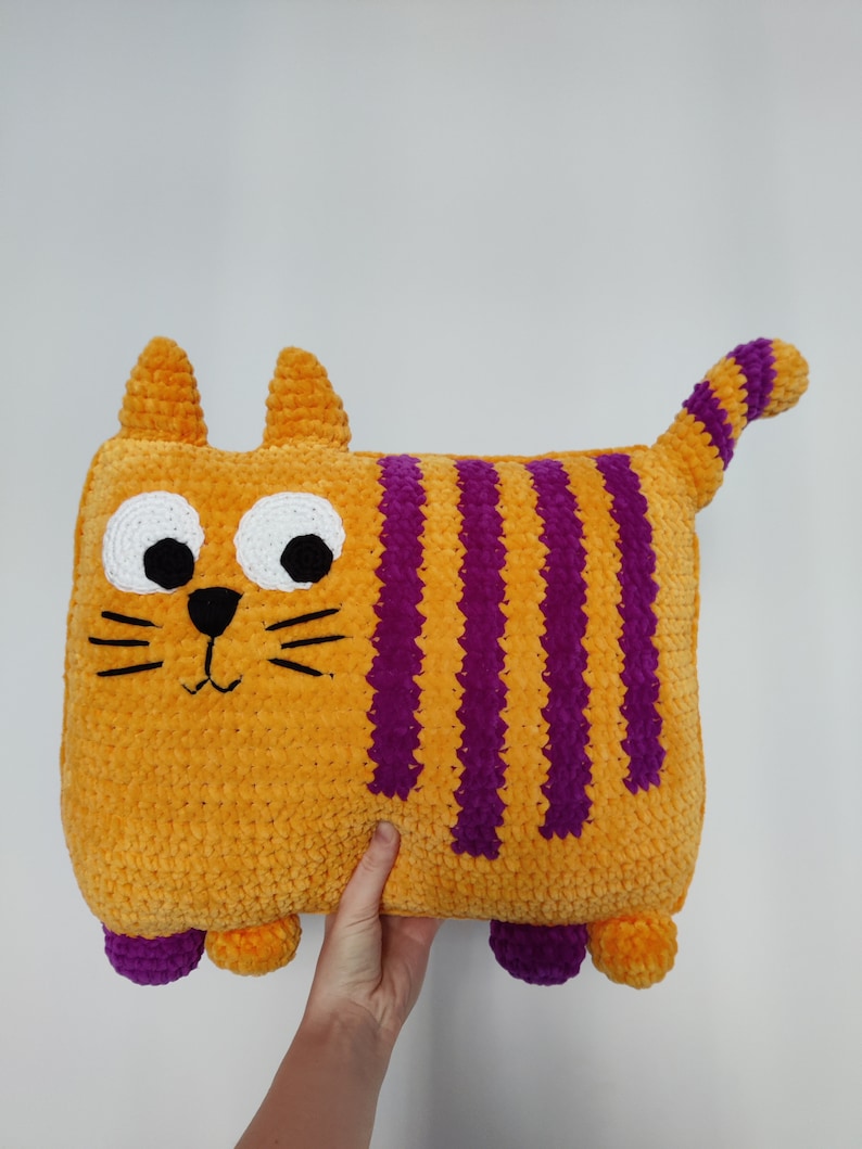May include: A handmade, orange cat-shaped cushion with purple stripes, white eyes, and a black nose and whiskers. The plush cushion has small orange ears, purple feet, and a striped tail. The cushion is held against a white background.