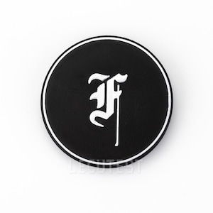 Può includere: Adesivo rotondo nero con una lettera "F" stilizzata bianca al centro.