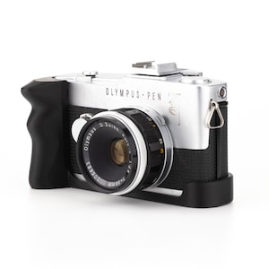 Puede incluir: Una cámara Olympus Pen F plateada con una empuñadura de cuero negro y una lente negra. La cámara tiene un lazo de correa plateado y el texto "OLYMPUS - PEN" está impreso en la parte superior.
