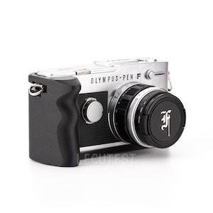 May include: A silver Olympus Pen F camera with a black lens cap. The camera has a black leather grip and a silver strap loop.