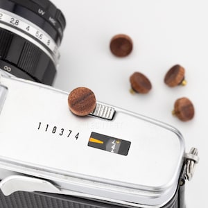 May include: A silver vintage camera with a black lens and wooden shutter release buttons. The camera has the number "118374" printed on it. Four wooden buttons are scattered around the camera.