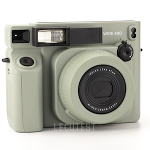 Op de afbeelding: Een lichtgroene instant camera met een groothoeklens en een ingebouwde flitser. De camera heeft een zwarte lensring met de tekst "INSTAX LENS 95MM" en "FOCUS RANGE 0.6M - ∞". De camera heeft ook een zwarte draaiknop met de tekst "WIDE 400" en een zelfontspannerknop.