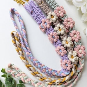 May include: Three colorful macrame keychains with flower accents. The keychains are made with a braided cord in shades of pink, blue, yellow, and brown. Each keychain has a gold key ring.