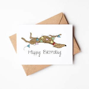 May include: A white greeting card with a brown dog illustration, wearing a red collar and a garland of colorful pennants. The card reads "Happy Birthday" in black script. The card is paired with a brown envelope.