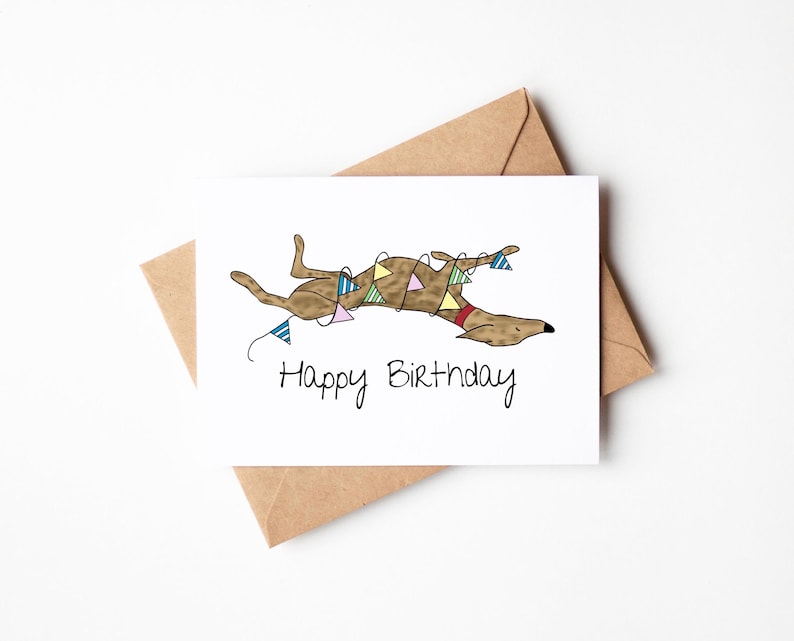 May include: A white greeting card with a brown dog illustration, wearing a red collar and a garland of colorful pennants. The card reads "Happy Birthday" in black script. The card is paired with a brown envelope.