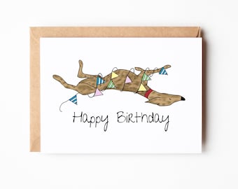 Può includere: Biglietto di auguri con un'illustrazione di un cane marrone, ornato di ghirlande e un collare rosso. Le parole "Happy Birthday" sono scritte in corsivo nero sotto il cane. Il biglietto è all'interno di una busta marrone.