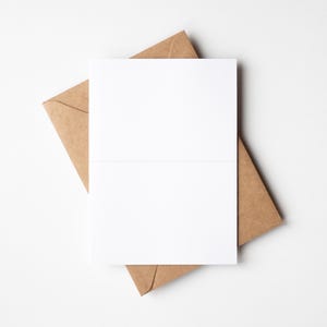 May include: A blank white folded card rests on top of a brown paper envelope. The card is centered, with the envelope's corner visible. The background is a plain white surface, creating a minimalist aesthetic.