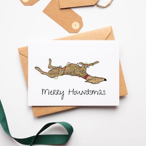 May include: A white greeting card featuring a brown dog illustration adorned with Christmas lights and a red collar. The card displays the text "Merry Houndsmas" in black script. The card rests on a brown envelope, with brown tags and green ribbon.