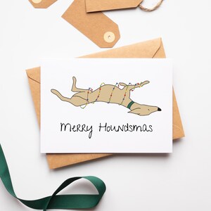 May include: A greeting card featuring a cartoon dog decorated with Christmas lights, lying on its back. The card reads "Merry Houndsmas" in black script. The card is on a brown envelope, with brown tags and green ribbon.