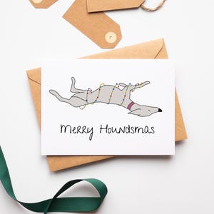 May include: A white greeting card featuring a cartoon greyhound dog decorated with Christmas lights, with the text "Merry Houndsmas". The card is on a brown envelope, with gift tags and a green ribbon.