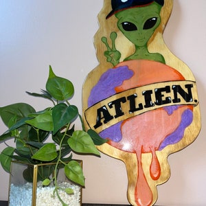 Könnte beinhalten: Ein grüner Außerirdischer mit einer blauen Baseballkappe mit dem Buchstaben "A" darauf, der ein Peace-Zeichen zeigt. Der Außerirdische hält einen schmelzenden orangefarbenen und violetten Globus mit dem Wort "ATLIEN" darauf.