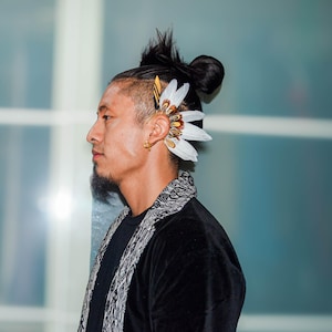 May include: A side profile of a person wearing a black top and a decorative ear cuff. The ear cuff features white feathers and gold accents. The person has dark hair styled in a bun and a patterned scarf.