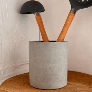 May include: A gray concrete utensil holder with a wooden base holds a black ladle and slotted spoon. The ladle and spoon have wooden handles. The kitchen tools are displayed on a round wooden cutting board.