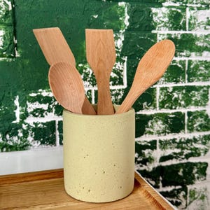 Op de afbeelding: Een lichtgroene cilindrische houder met vier houten keukengerei. Het keukengerei omvat een lepel en drie spatels. De houder staat op een houten dienblad, met een groen en witte bakstenen muur op de achtergrond.