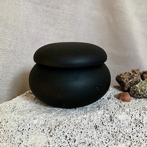 May include: A small, black, round wooden container with a lid. The container is sitting on a white, textured surface. There are small, colourful stones around the container.