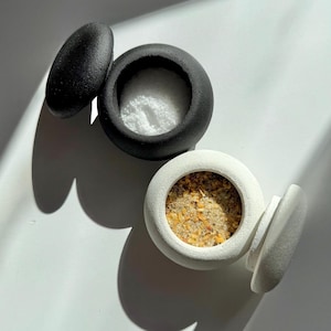 Può includere: Due piccoli contenitori rotondi in ceramica con coperchi. Uno è nero e riempito di polvere bianca, l'altro è bianco e contiene un mix di spezie. I coperchi sono ovali e si trovano accanto ai contenitori.