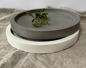 May include: Two round concrete trays, one gray and one white, stacked on top of each other. A small sprig of greenery rests on the gray tray. The trays have a minimalist design and a smooth texture, suitable for home decor.