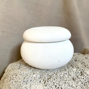 Puede incluir: Un pequeño recipiente de cerámica blanco con un cuerpo redondeado y una tapa a juego. El recipiente tiene una superficie texturizada y se asienta sobre una piedra rugosa de color claro. El fondo es una tela neutra blanquecina.