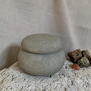May include: A gray stone container with a lid. The container is round and has a rough texture. It is sitting on a white, textured surface.