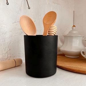 May include: A black cylindrical utensil holder with wooden spoons and a honey dipper inside. The spoons are light brown and have different shapes. A rolling pin is on the left. A white teapot is in the background.
