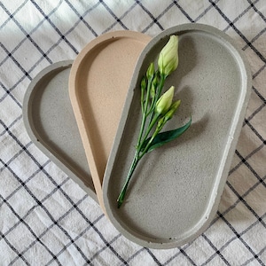 Puede incluir: Dos bandejas de hormigón ovaladas, una gris y otra beige, con una rama de flores blancas encima de la bandeja gris.