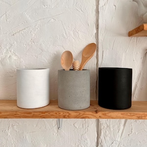 Puede incluir: Tres soportes para utensilios cilíndricos de hormigón en blanco, gris y negro en un estante de madera. Dos cucharas de madera están en el soporte gris.