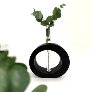 May include: A black circular vase with a glass test tube in the center, holding a sprig of green eucalyptus.
