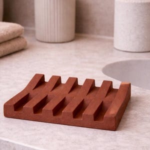Peut inclure: Un porte-savon rectangulaire brun rougeâtre avec un design à fentes. Le porte-savon est fait d'un matériau solide et repose sur un comptoir de couleur claire. D'autres accessoires de salle de bain sont visibles en arrière-plan.