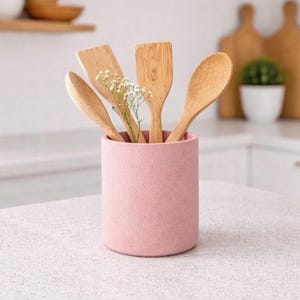 May include: A pink speckled utensil holder containing wooden spoons and spatulas. The holder has a small sprig of white flowers. The kitchen utensils are made of light-coloured wood. The background includes a white countertop and white cabinets.
