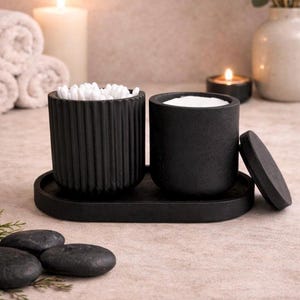 May include: A set of two matte black cylindrical containers on an oval tray. One container has vertical ribbed detailing and holds white cotton swabs. The other container has a smooth surface, contains white cotton pads, and has a matching lid.