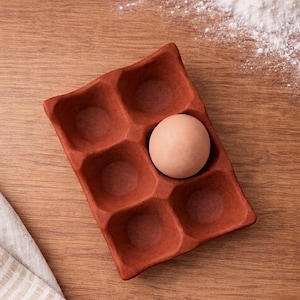 Op de afbeelding: Een terracotta-kleurige eierhouder met zes compartimenten, waarvan één met een enkel bruin ei. De houder staat op een houten oppervlak, met een linnen doek en een snufje bloem op de achtergrond. Een rustiek keukenartikel.