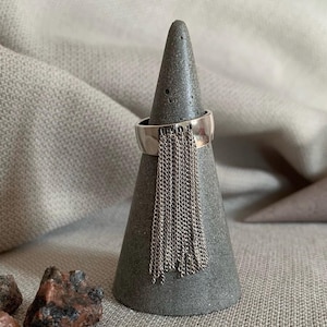 Può includere: Un anello d'argento con una fascia larga e una frangia di catenelle pendenti. L'anello è esposto su un porta anelli a forma di cono in cemento grigio. Lo sfondo è un tessuto neutro e piccole rocce sono in primo piano.