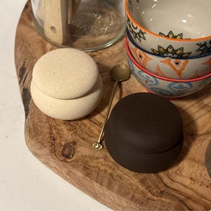 May include: A wooden serving board displays a beige, round container, a dark brown round container, a small gold spoon, and two decorative bowls with orange and blue patterns. The containers appear to be ceramic.