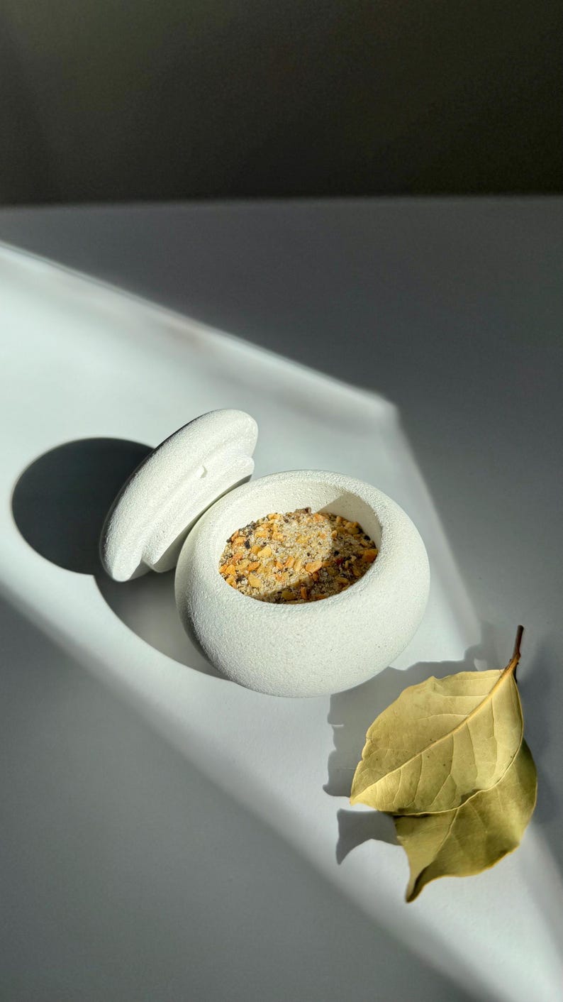 Op de afbeelding: Een witte keramische container met een deksel, gevuld met een mengsel van witte en bruine specerijen. Een enkel geel blad is op de achtergrond.