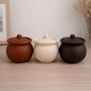 May include: Three small, round ceramic jars with lids. The jars are in shades of brown, cream, and dark brown, arranged side by side on a wooden surface. Each jar has a small, round knob on top of the lid.