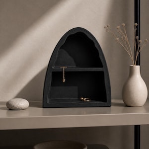 May include: A black, arched jewelry organizer with two compartments. A gold necklace and ring are displayed inside. A small, beige vase with dried flowers and a smooth, gray stone are on the shelf. A small, beige bowl is on the shelf below.